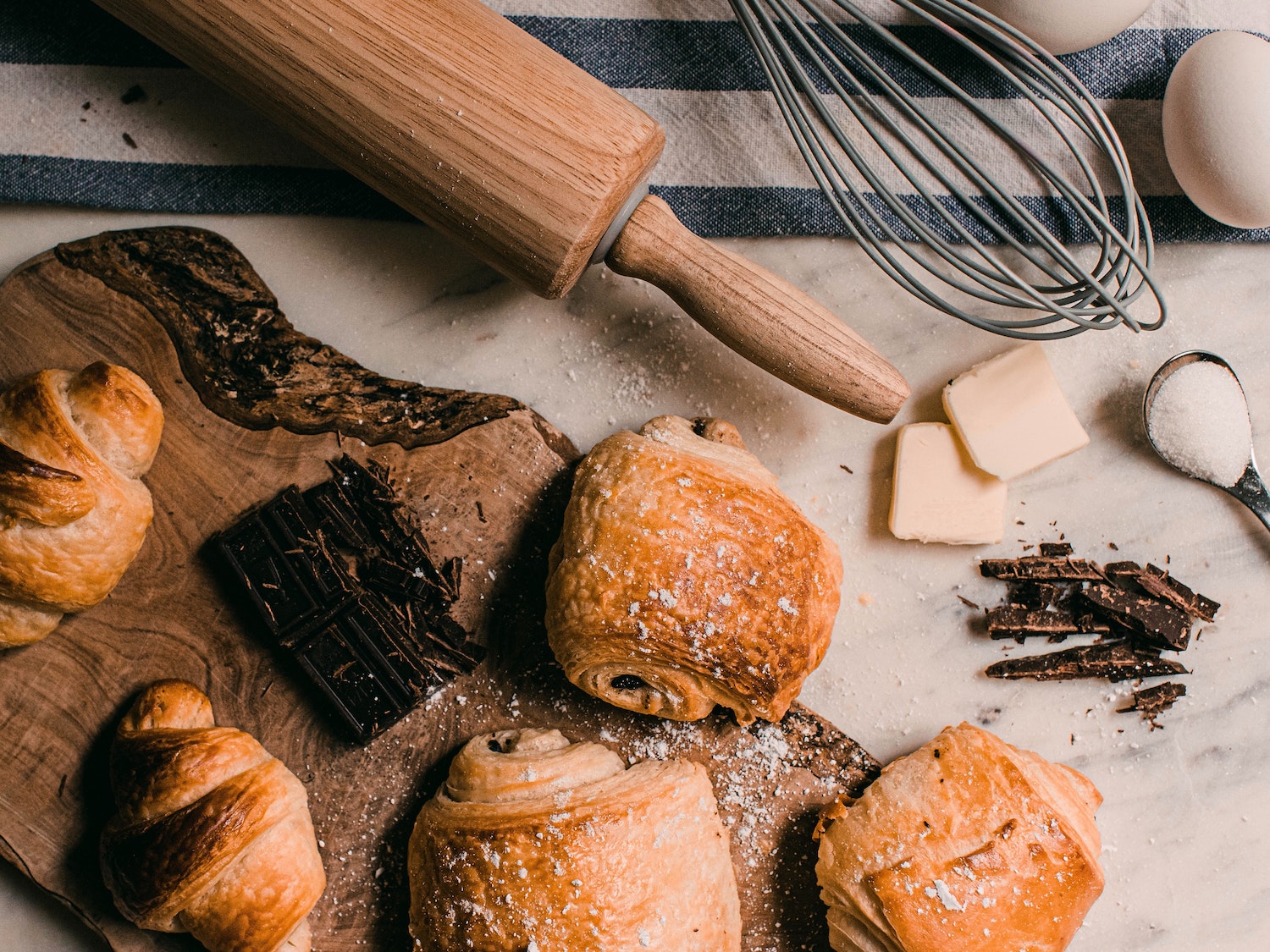 Several pastries on a wooden board beside a rolling pin, whisk, and ingredients